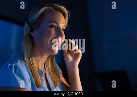 Female customer representative communicating with customer through headset in office at night Stock Photo