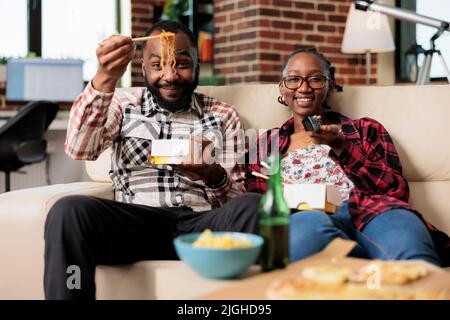 Happy couple watching television and eating noodles meal, switching tv channel program with remote control. Having takeaway dinner food while they watch movie or film, leisure activity. Stock Photo