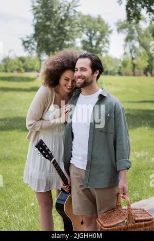 man in summer clothes carrying guitar and wicker basket near cheerful girlfriend in green park Stock Photo