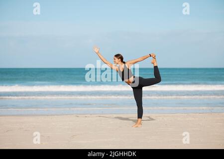 This pose develops greater balance and core strength. a young woman holding a dancer pose while practising yoga at the beach. Stock Photo