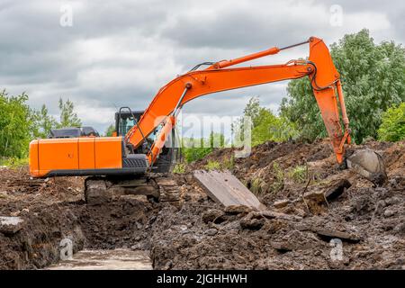 Large orange crawler excavator is excavating (digging a trench) against backdrop of green trees and an overcast dramatic summer sky. Stock Photo