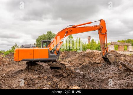 Large orange crawler excavator is excavating (digging a trench) against backdrop of green trees and an overcast dramatic summer sky. Stock Photo