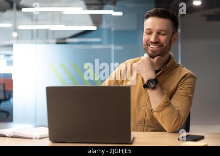Cheerful Business Man Using Laptop Touching Chin Sitting At Workplace Stock Photo