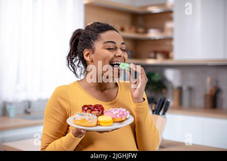 Glad millennial pregnant african american woman hold plate of sweets and eating cookies Stock Photo