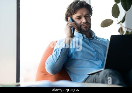 Handsome mature businessman talking on cellphone, using laptop, working in modern office, free space Stock Photo