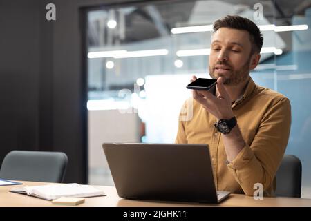 Businessman Using Voice Search On Cellphone Working On Laptop Indoor Stock Photo