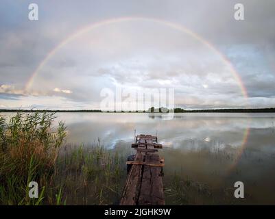 An old wooden bridge on the lake on the background of a beautiful rainbow in a cloudy sky. Landscapes the hinterlands of russia Stock Photo