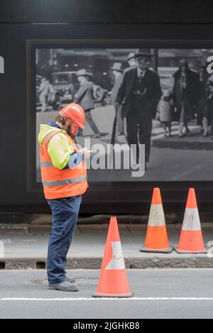 A man stands in Sydney wearing high vis clothing and a hard hat, looking at his phone. In the background an old photo of a man in a suit and hat Stock Photo