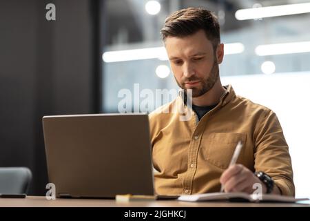 Serious Businessman Using Laptop Computer Taking Notes In Modern Office Stock Photo