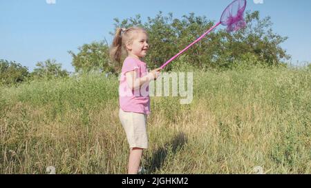 Little girl plays with butterfly net of tall grass in city park. Cute little girl is playing with aerial insect net in meadow on sun day. Stock Photo