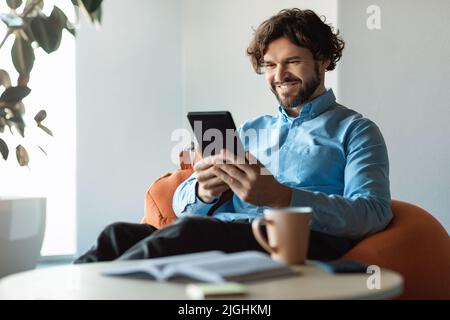 Happy mature businessman using tablet pc, looking at screen, sitting on bean bag chair in modern office, empty space Stock Photo