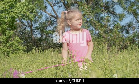 Little girl plays with butterfly net of tall grass in city park. Cute little girl is playing with aerial insect net in meadow on sun day. Stock Photo