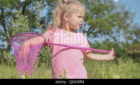 Little girl plays with butterfly net of tall grass in city park. Cute little girl is playing with aerial insect net in meadow on sun day. Stock Photo