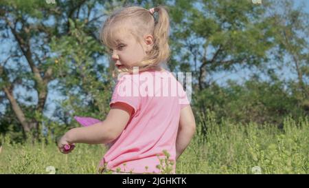 Little girl plays with butterfly net of tall grass in city park. Cute little girl is playing with aerial insect net in meadow on sun day. Stock Photo