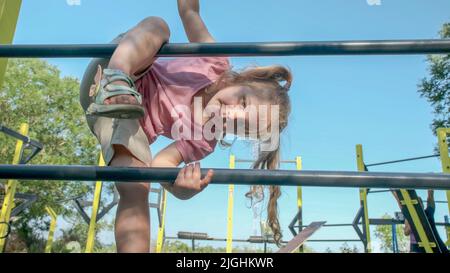 Little girl climbs gymnastic ladder on open sports ground on outside. Cute little girl crawls on vertical sports ladder in city park on sun day. Stock Photo