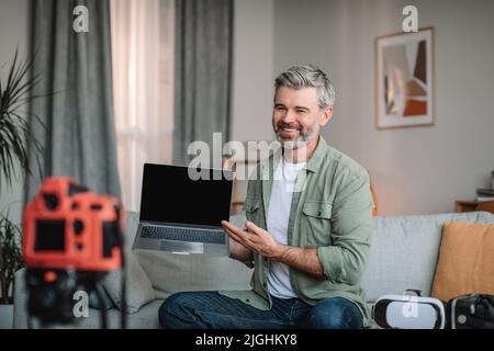 Cheerful european old man with beard shows laptop with blank screen, shoots video in room interior Stock Photo