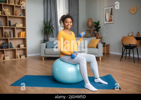 Cheerful young pregnant african american female with big belly doing exercises with dumbbells on fit ball Stock Photo