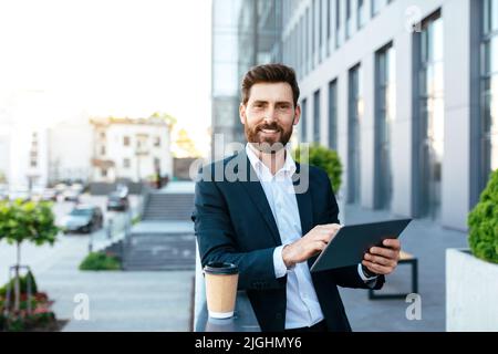 Smiling handsome confident european millennial bearded businessman in suit uses tablet to work Stock Photo