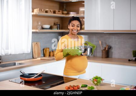Smiling young pregnant african american woman with big belly in protective gloves prepares dinner Stock Photo