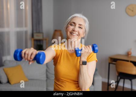 Positive senior woman doing dumbbell workout at home, working on arms strength and smiling at camera Stock Photo