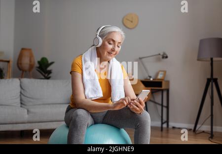 Senior woman using smartphone and wearing headphones, sitting on fitball during online domestic workout Stock Photo