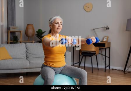 Active retirement. Happy senior woman in sportswear exercising with dumbbells on fitball, having domestic training Stock Photo