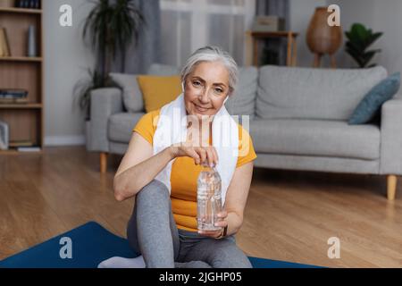 Senior woman in sportswear sitting on sports mat with bottle of clear water, keeping hydrated during domestic training Stock Photo