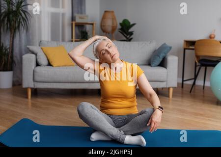 Healthy lifestyle concept. Senior woman stretching neck, sitting on yoga mat and warming up before domestic workout Stock Photo