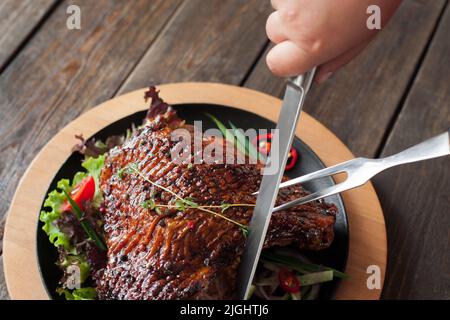 Cutting grilled duck thigh close-up top view. Hands carving of roasted poultry leg with two-pronged fork and knife on wooden background Stock Photo