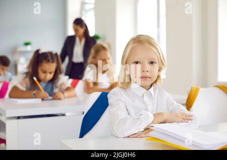 Portrait of cute little serious boy sitting at desk during lesson in school classroom. Stock Photo
