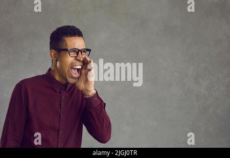 African american man stands on a gray background and shouts holding his hand to his mouth. Stock Photo