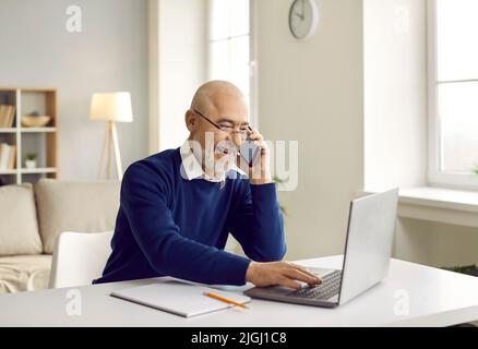 Happy senior man sitting at his desk, talking on his mobile phone and using his laptop Stock Photo