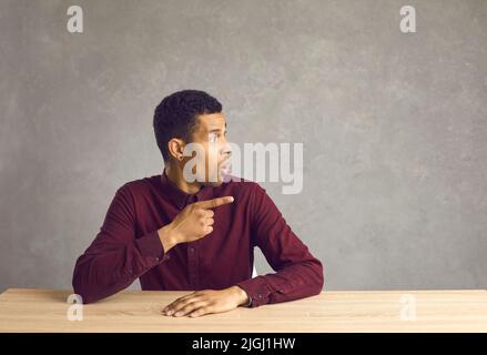 Surprised young black man sitting at desk looking and pointing away at copy space background Stock Photo