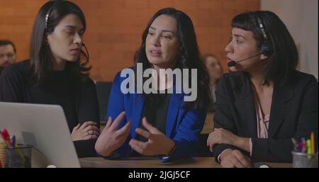 Business woman coaching younger staff in front of computer laptop. Female leader explaining protocol to staff wearing headsets at office Stock Photo