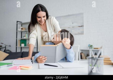 nanny smiling near child sitting with textbook near copy books on table Stock Photo