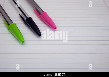Close-up of three pens with caps on resting on a lined paper sheet. Background of green, black and pink pens with selective focus and empty space Stock Photo