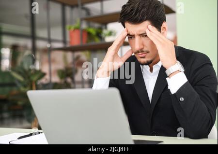 Fatigued hispanic businessman using laptop for working, the guy tired and burnout, holding head and feels headache, migraine sitting at the desk in modern office. Overwork and burnout concept Stock Photo