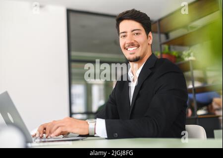 Optimistic confident dark-haired latin handsome male employee wearing formal suit using laptop for work in the office, businessman sits at the desk and looks at camera, smiles friendly Stock Photo