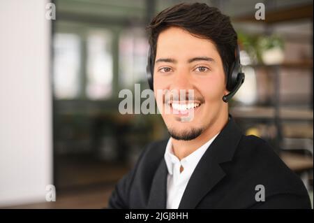 Male support worker in touch. Video chat with young smiling latin man wearing headset looks at camera, talks with customers or colleagues on distance, male employee involved video meeting, webcam view Stock Photo