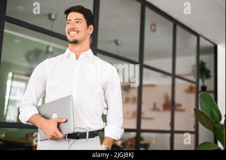 High-skilled smiling young businessman carrying laptop in a modern office, white collar worker ready for new working day, inspired with new ideas male start-up owner looking aside Stock Photo