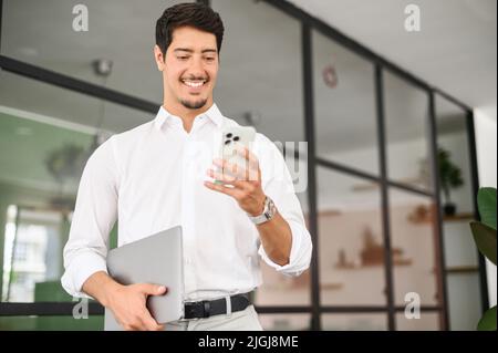 Handsome successful hispanic male entrepreneur using smartphone indoors, walking through hall and carrying laptop, smiling businessman texting on the phone Stock Photo