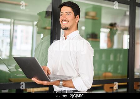 Smart and intelligent developer guy is working with laptop standing in contemporary coworking space. Ambitious young latin man in smart casual white shirt holding laptop and looks aside lost in dreams Stock Photo