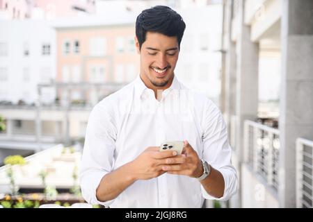 Handsome hispanic guy is using smartphone for messaging, texting, young dark-haired man in smart casual white shirt using smartphone outdoors, typing a message, chatting in the networks, scrolling Stock Photo
