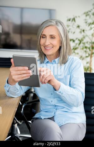 Vertical portrait of mature businesswoman sit in office and using digital tablet Stock Photo