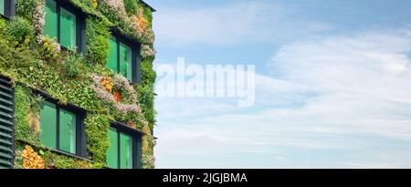 Exterior of a green sustainable building covered with blooming vertical hanging plants in front of a blue sky Stock Photo