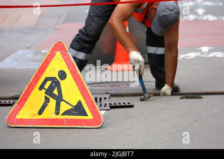 Road works sign, worker of municipal services repairs drainage system. Wastewater treatment, orange roadblock cones on the city street Stock Photo