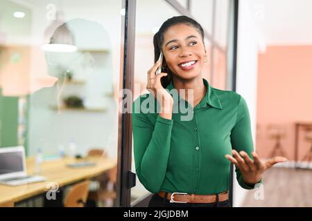 Friendly smiling african-american female employee has pleasant phone conversation standing in modern office space, cheerful black businesswoman in green shirt talking on smartphone and gesturing Stock Photo