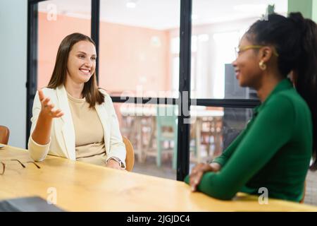 Two young happy multiracial female colleague sitting at desk and communicating while working together in coworking space, sharing new ideas, discussing project strategy Stock Photo