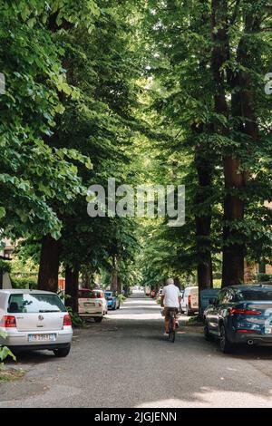 Venice, Italy - May 22, 2022: Rear view of a man cycling past parked cars on a tree-lined street on Lido Island, a barrier island in Venetian Lagoon f Stock Photo