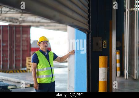 Asian warehouse worker in coveralls and protective helmet opening shutter roller door at the beginning of working day Stock Photo